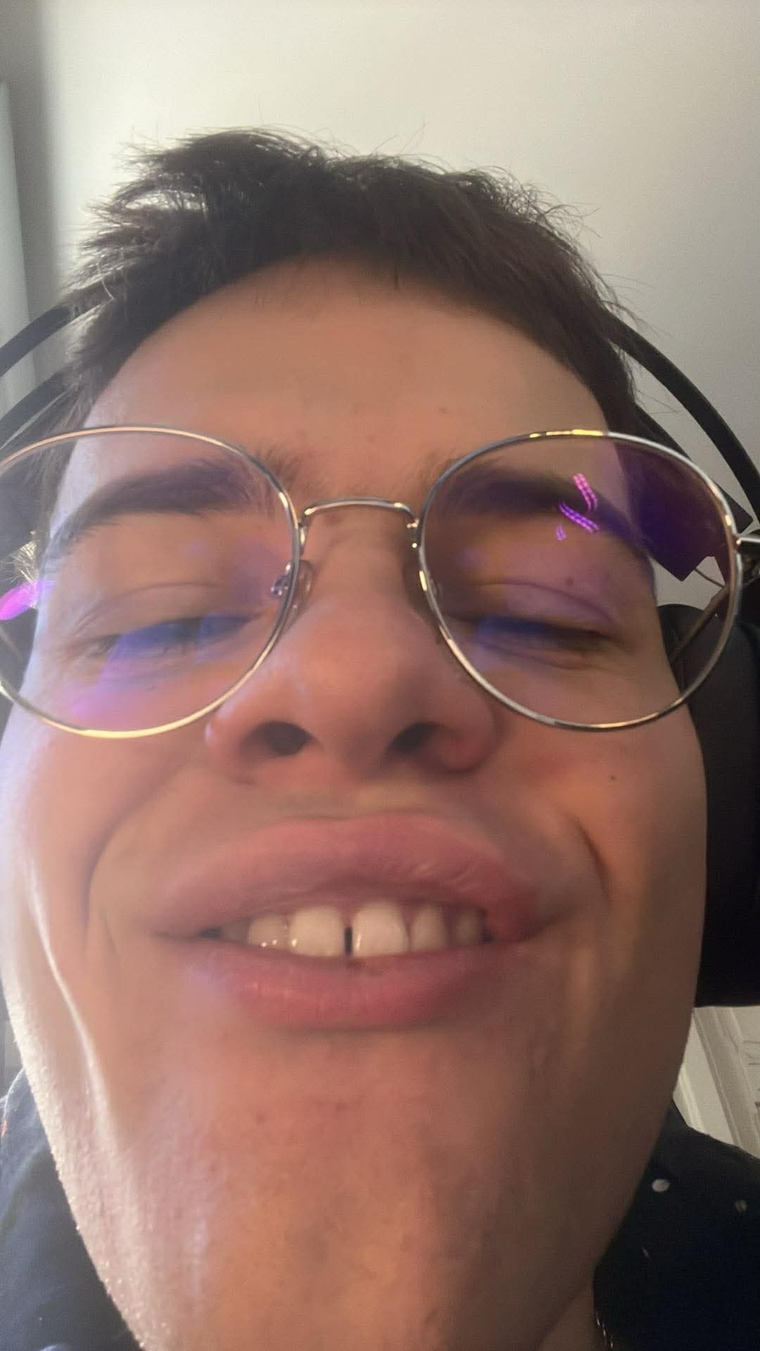 Close-up, low-angle selfie of person wearing round glasses and headphones, smiling with a gap-toothed grin.